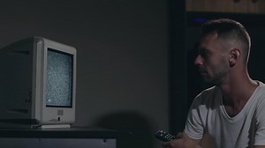 Broken Television or TV Propaganda. Mind Control - Zombified Man in White T-shirt Switches Channels on the Old TV in Dark Room. TV Signal Search. Distortion on the Screen. Medium Close-Up, Film Grain
