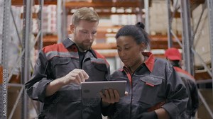 Medium shot of caucasian construction contractor explaining working plan to african-american female working person both wearing coveralls and looking at tablet standing at big warehouse