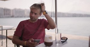 Male tourist having drink at a cafe in the city near sea. Man on a coffee break checking the phone. 50fps slow motion.