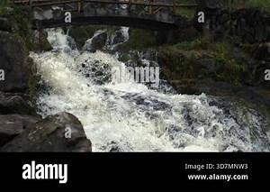 Waterfall cascading beneath a wooden bridge over dark rocks and mossy terrain in Ultra slow motion captured with a high-speed camera