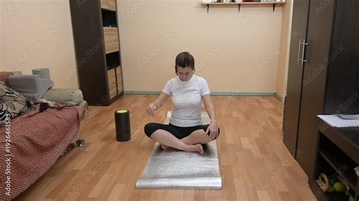 Young woman sitting on a yoga mat at home and doing neck stretching exercises to relieve tension