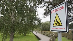 Caution slippery floor sign in Spanish, person ride bike on park path in background