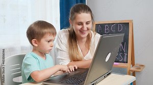 Smiling mother teaching her little son using laptop computer during education and studying at home. Concept of remote school and distance education at home