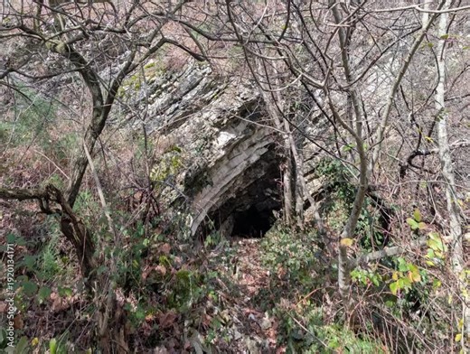 ingresso di una grotta naturale all’interno di una formazione rocciosa stratificata. Le rocce inclinate e fratturate evidenziano processi geologici antichi