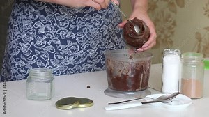 A woman puts nut butter from a blender bowl into a jar. Walnut urbech. The ingredients are on the table side by side