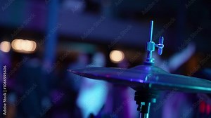 The stage is prepared with a drum set, cymbals, and musical instruments under a blue spotlight, creating an exciting ambiance for a concert. The setting is dynamic and full of energy