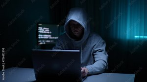 Front view of stealthy hacker man wearing sweatshirt with hood typing on keyboard breaking password using laptop, sitting at desk in dark room, computer monitor with text code on screen on background.