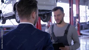auto service, mechanic explaining expenses for car repair to his client writes in clipboard and shaking hands on background the automobile on lift