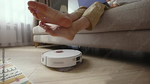 Robot vacuum cleaner driving around barefoot woman at home. Low angle shot of contemporary robotic vacuum cleaner working on floor beside female feet. Electrical appliances modern technology concept