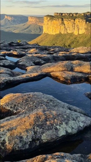 A Chapada Diamantina Fica na Bahia em primeiro plano formações rochosas areníticas, erosionadas ao longo de milhões de anos, apresentam piscinas naturais que refletem o céu. Ao fundo, imensos paredões de arenito e conglomerados se erguem, revelando camadas de história geológica e a vastidão da paisagem. A vegetação exuberante de caatinga e cerrado cobre as encostas, enquanto a luz do sol poente doura as formações rochosas, criando um espetáculo visual inesquecível. Esta imagem nos convida a expl