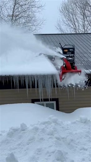 The footage opens on a residential neighborhood buried under heavy snow, where a man stands on a rooftop clearing thick drifts after a long storm. The snow blower roars loudly as it chews through packed ice, sending white powder flying into the air. Everything appears routine until the machine suddenly jerks and shifts direction. In a split second, the snow blower surges forward uncontrollably, dragging the man across the slick surface. He struggles to regain footing as the machine pulls him tow