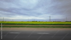 Meadows fields, electric power transmission towers on background. Highway with cloudy weather, overcast. View through side window of van in motion. Roadway out of town