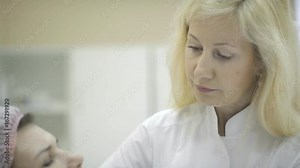 Experienced cosmetologist talks to patient, examining her face indoors. She carefully surveys skin of adult woman sitting in front of her and speaks about details of upcoming treatment. Skillful