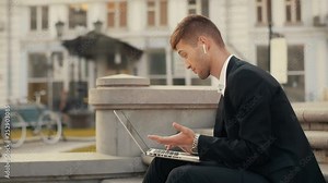 Young man in suite working on laptop holding it on the lap. Disappointed businessman with wireless headphones working out of office sitting at the city square on stairs with bicycle on the background