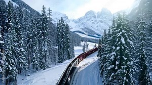 A train winds through a stunning winter landscape, surrounded by towering snow-covered mountains, with early morning light casting a soft glow across pristine snow.