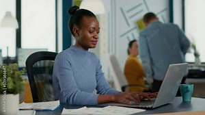 Portrait of african american woman typing using laptop keyboard and smiling at camera sitting at desk in busy startup office. Casual business employee working relaxed with sales statistics.