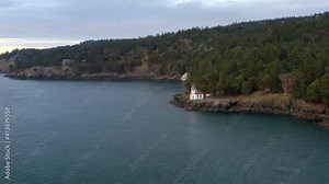 Drone flying away from lighthouse at sunset in the Pacific North West San Juan Islands. Birds flying over ocean with forest in background. Lighthouse