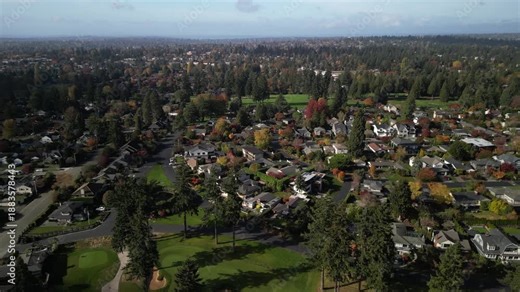 Aerial drone view of an oval-shaped residential neighborhood surrounded by a golf course in Seattle. The drone flies laterally from left to right, revealing homes, fairways, and colorful fall foliage.