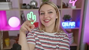 Victory! young blonde female streamer, flashing a cheerful v sign, winning at her game. smiling happily at the camera in a dark gaming room.