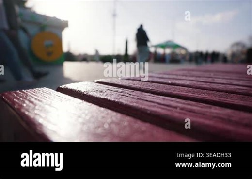 Dark red painted wooden planks form the seat of a public park bench. Blurred pedestrians walk past in the bright background. The shallow depth of field highlights the textured wood Stock Video Footage - Alamy