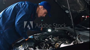 Professional auto mechanic examining car engine, looking under hood with flashlight, side view
