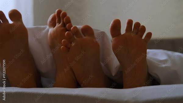 Loving couple play and dance their feet under white blanket while wake up in bed in morning. Young man and woman feet under sheets on the bed at home. Close up view