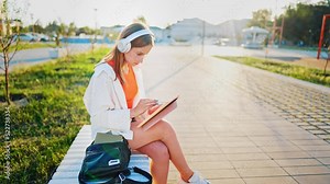 Teenage girl with headphones sitting on park bench and drawing with stylus on graphics tablet. Artist is engaged hobby in nature. Businesswoman with tablet laptop at work outdoors. Freelance online.