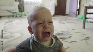 Cute baby crying bitterly, caucasian baby with blonde hair sitting on the floor, toddler upset and roaring opening his mouth wide and baring his teeth.