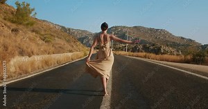Happy young girl in a light brown dress walk gentle barefoot into the distance on a empty country road in beautiful landscape. Lovers road trip adventures concept