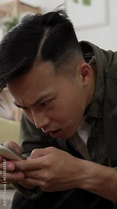 Vertical Video: closeup view of a confused asian man finger pointing at the screen while checking a surprising notification message on the smartphone in the living room at home