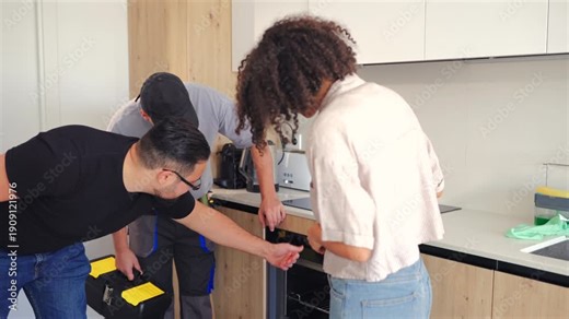 Professional handyman repairs kitchen appliance in modern apartment while tenants observe. Indoor maintenance service, home improvement work and property management concept.