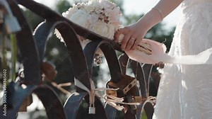 Elegant bride with wedding bouquet standing near the steel gate with closed locks as a symbol of forever love, wind waving flower bouquet ribbon and bridal veil, charming wedding details of bride
