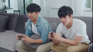 Young Asian gay couple play games at home, Teen korean LGBTQ men using joystick having funny happy moment together on sofa in living room at house. Slow motion shot.