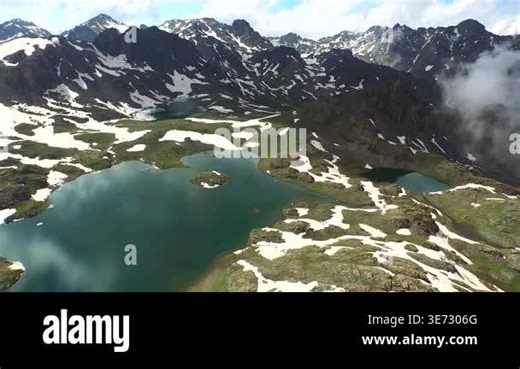 Aerial view of alpine mountain lake surrounded by melting spring snow and rugged peaks. High elevation tundra and green meadow landscape create breathtaking remote wilderness scenery Stock Video Footage - Alamy
