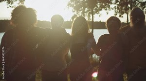 backlit silhouette of a group of young adults hugging in a row walking together in an idyllic natural setting at sunset among trees and lake