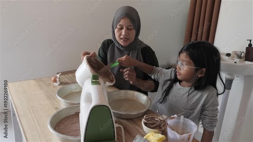Asian muslim mother making cake with her daughter, family cooking together, pouring batter in baking pan