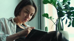 Woman artist drawing on digital tablet. Creative girl holding notepad with digital pencil in hands and working remotely from home. Freelancer using computer to create art sitting near window