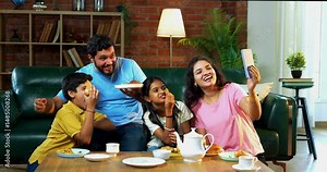 Video calling while eating samosa and drinking tea at home, Indian parents with kids bonding together, chatting with friends or relatives, enjoying snacks in cozy modern living room setting