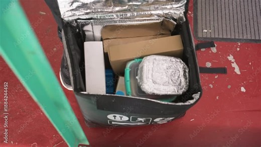 courier packing insulated delivery bag with meals. hands place meal containers, cardboard boxes and cooler pack inside reflective liner red floor and green door frame signal urban storefront