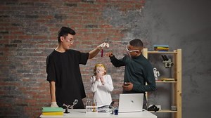 Joyful Asian and African-American chemists hold test tube above girl student shooting video for blog at online fun science lesson in improvised classroom