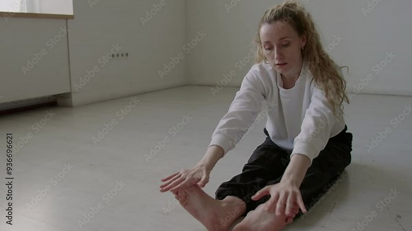 Young ballerina sitting on floor, practicing forward bend with toe touch and flexing feet during stretching in studio