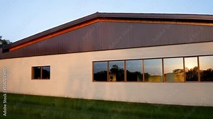 A modern farmhouse with soft beige siding and a dark brown metal roof, set in a lush, grassy field. Large windows bring a contemporary touch to the rustic charm of the house.