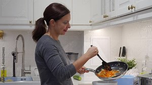 Adult 44 year old woman doing her daily cooking in the kitchen at home. Woman puts fried vegetables from a frying pan into a bowl.
