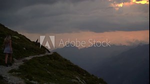 Very cinematic footage of a blonde girl in a white dress walking on an exposed mountain path towards the rising sun until she disappears behind the mountain. It looks like a movie scene.