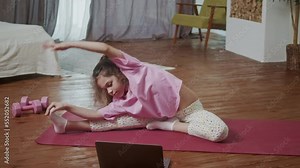 Little girl doing stretching exercises, practicing yoga on fitness mat at home in front of open laptop. Teenager kid doing horizontal scale workout looking laptop in light room interior.
