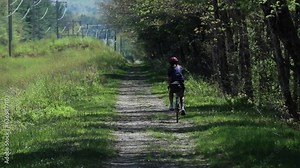 woman biking on a gravel path in the woods (bikepacking, bike touring, wooded area, rail trail, mountain riding) jersey, helmet, road cycling (shot from behind) singletrack, trees, grass
