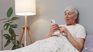 Senior guy enjoying digital interaction. Smartphone usage in the morning for the elderly. Caucasian elderly man in sleeping mask lying on bed using mobile phone at home