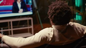 Man holding remote, watching news channel on television broadcast. Focus on person relaxing on couch in apartment by looking at widescreen smart TV set in blurry background