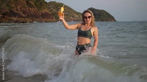 Woman stand knee-deep in sea on sunset. Girl on beach in green swimsuit having fun, drinks her orange cocktail, big waves hitting legs