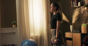 Deep squat. Side view of a young beautiful woman in sportswear doing a squat and holding dumbbells while standing in front of the window listening to music on headphones.